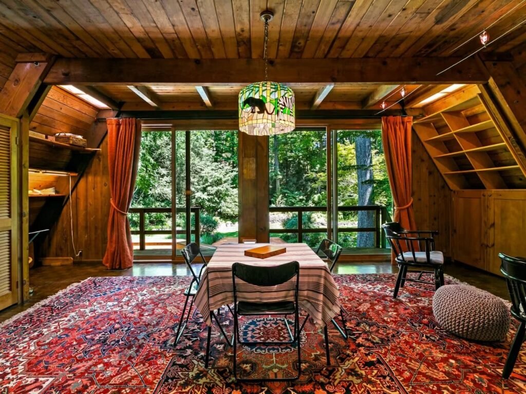 Interior of the open-plan dining area at the Woods Home. A dining table with a striped cloth and black chairs sits in front of expansive, floor-to-ceiling glass sliding doors that open onto a deck. Large trees in the forest are visible outside. The distinctive Tiffany-style lamp hangs above.