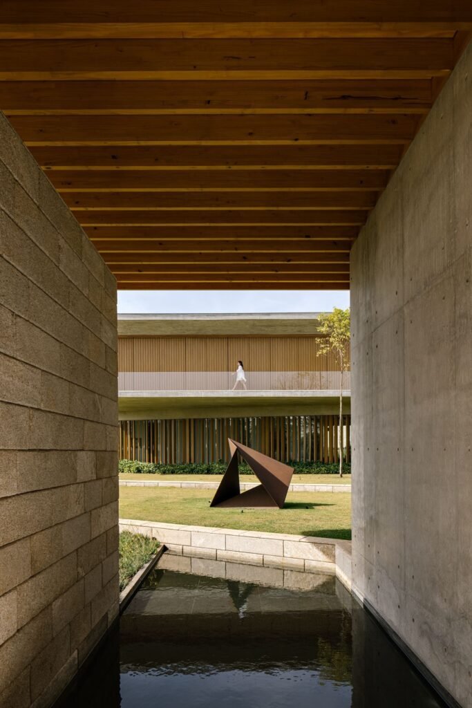 View through a stone and concrete passage across a reflective water feature toward an outdoor sculpture.