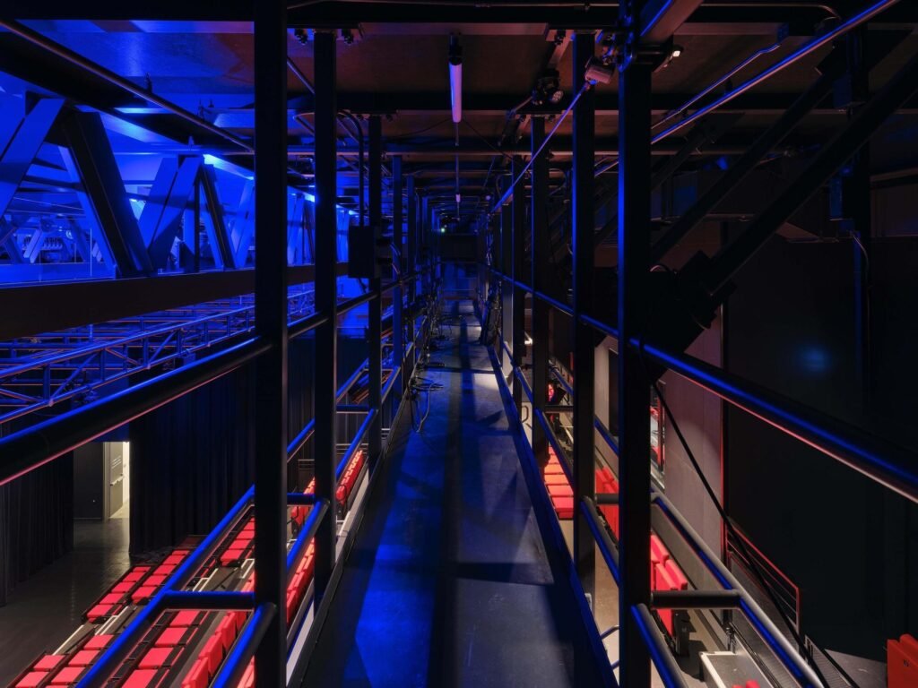 High-angle technical catwalk view inside the black box theater showing steel trusses and overhead blue lighting grid.