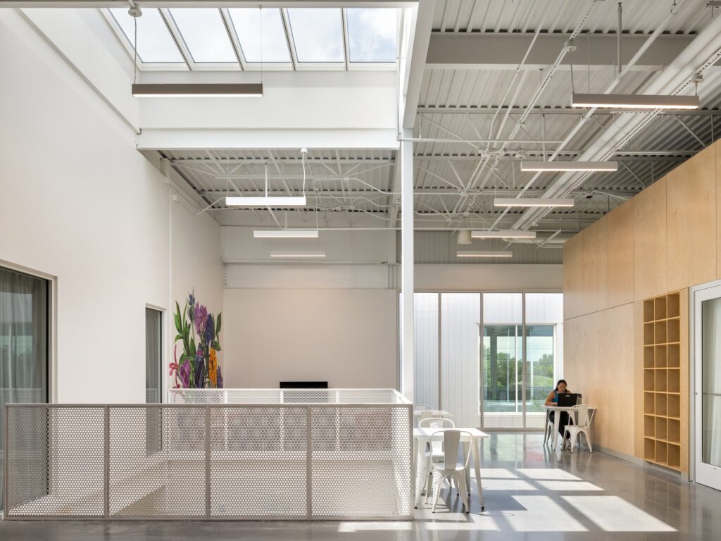 Bright mezzanine lounge level with work tables, linear skylights, exposed ceiling trusses, and perforated railings.