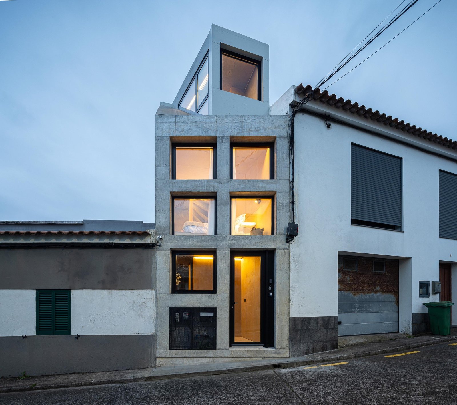 Evening view of Mini Atalhada facade with warm interior lights glowing through large glass windows framed by concrete structure.