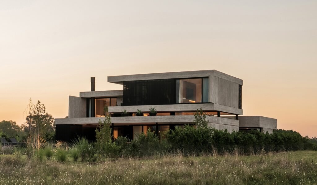 Exterior dusk view of concrete house showing illuminated interior lights against sunset sky and tall prairie grass.