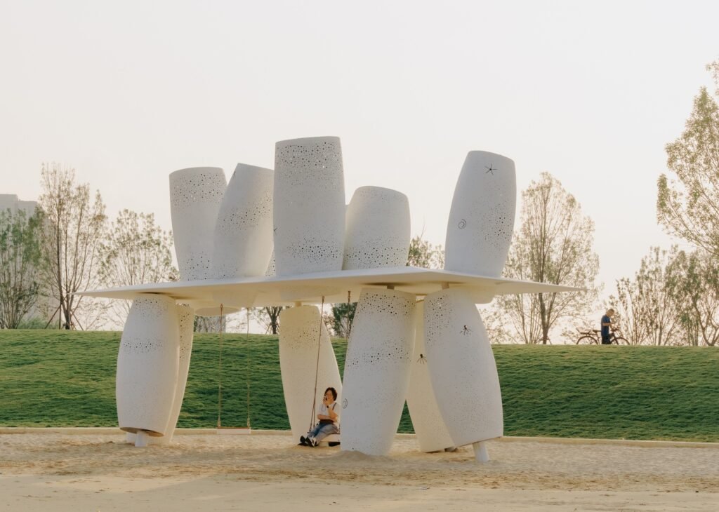 Eye-level view of the white Coral installation framed against a grassy hill, mature trees, and a clear sky with a visitor sitting on a swing.