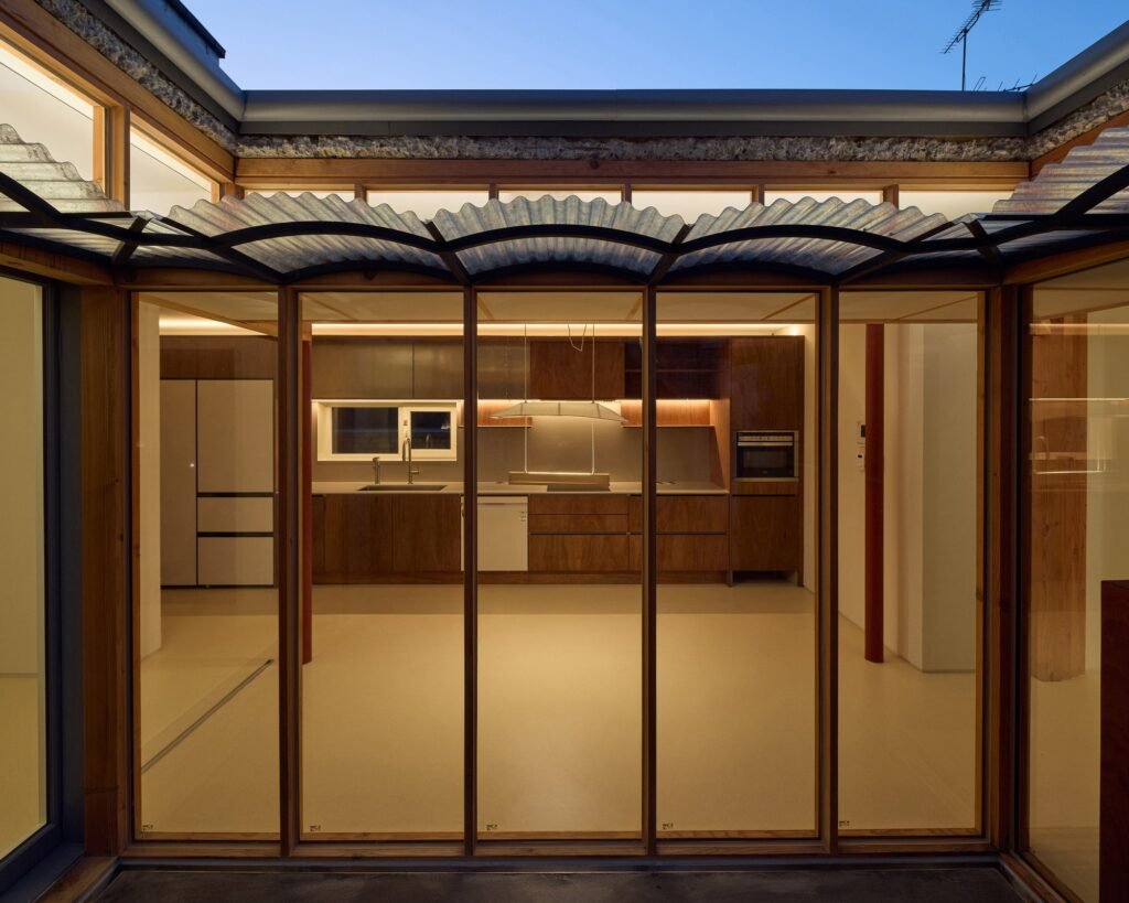 Evening view from the central courtyard looking through floor-to-ceiling glass doors into the warm timber-paneled kitchen of Saekdong House.