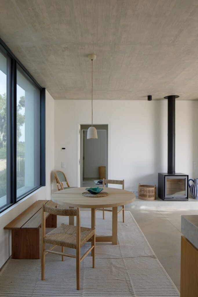Dining nook in Melides MG House with round wooden table, woven chairs, concrete ceiling, and modern wood stove.