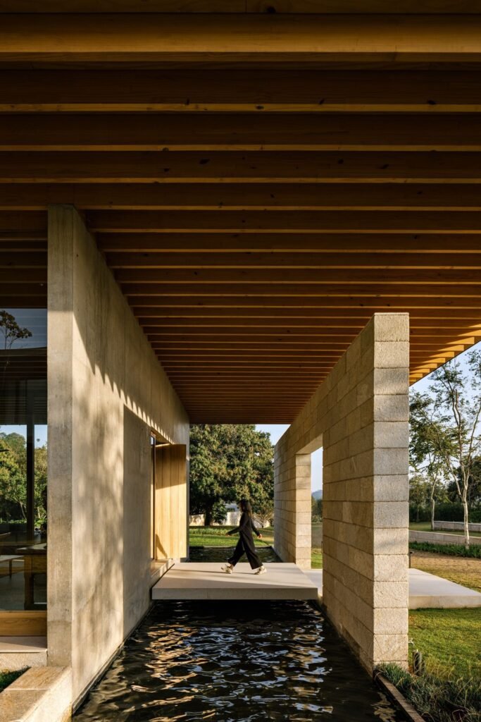 Covered walkway between stone wall and concrete facade with exposed wooden ceiling beams and a reflective water channel.