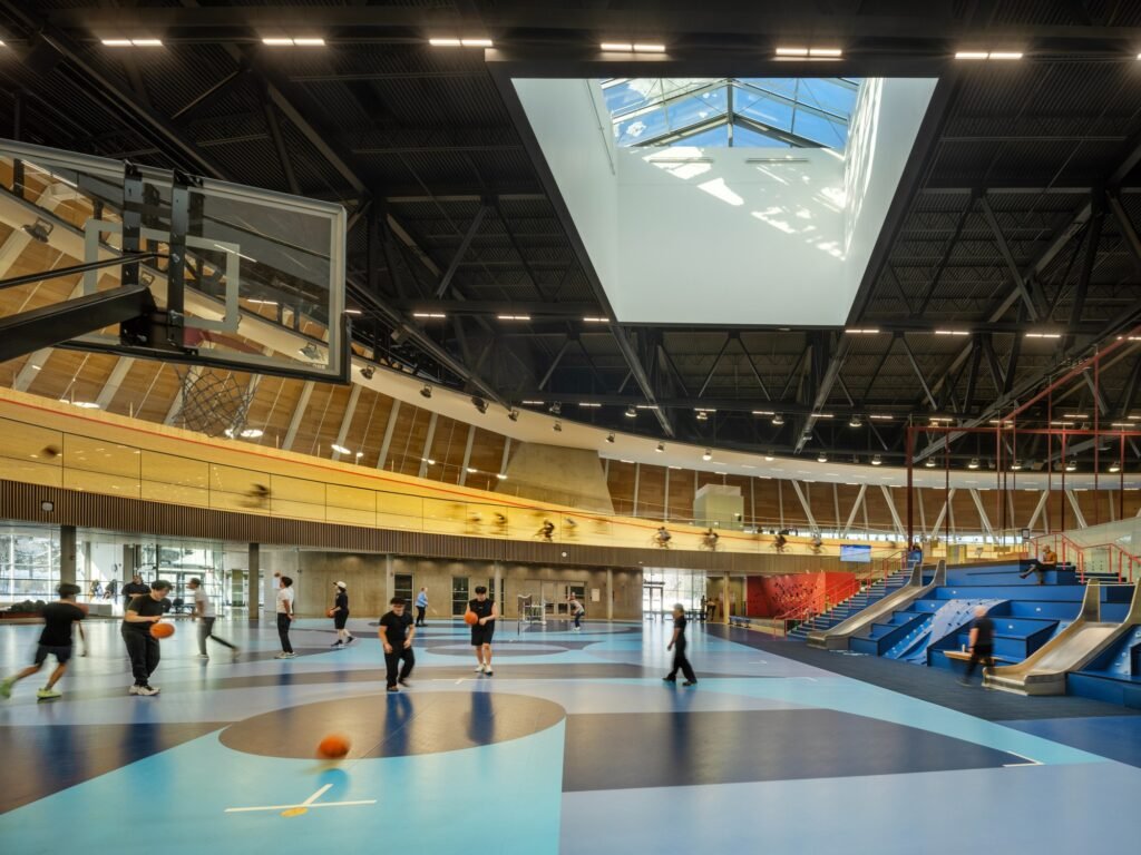 Wide interior view looking across the ground-floor sports court towards the elevated cycling track visible above through transparent barriers.