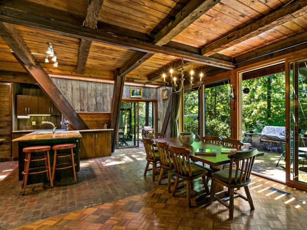 An interior view of the Woods Home open-plan kitchen and dining space. A long wooden dining table with a traditional brass chandelier hangs above. To the left is the kitchen with an island, stone countertops, wooden cabinets, and bar stools. A brick floor section transitions to wood. More glass doors show the forest outside.