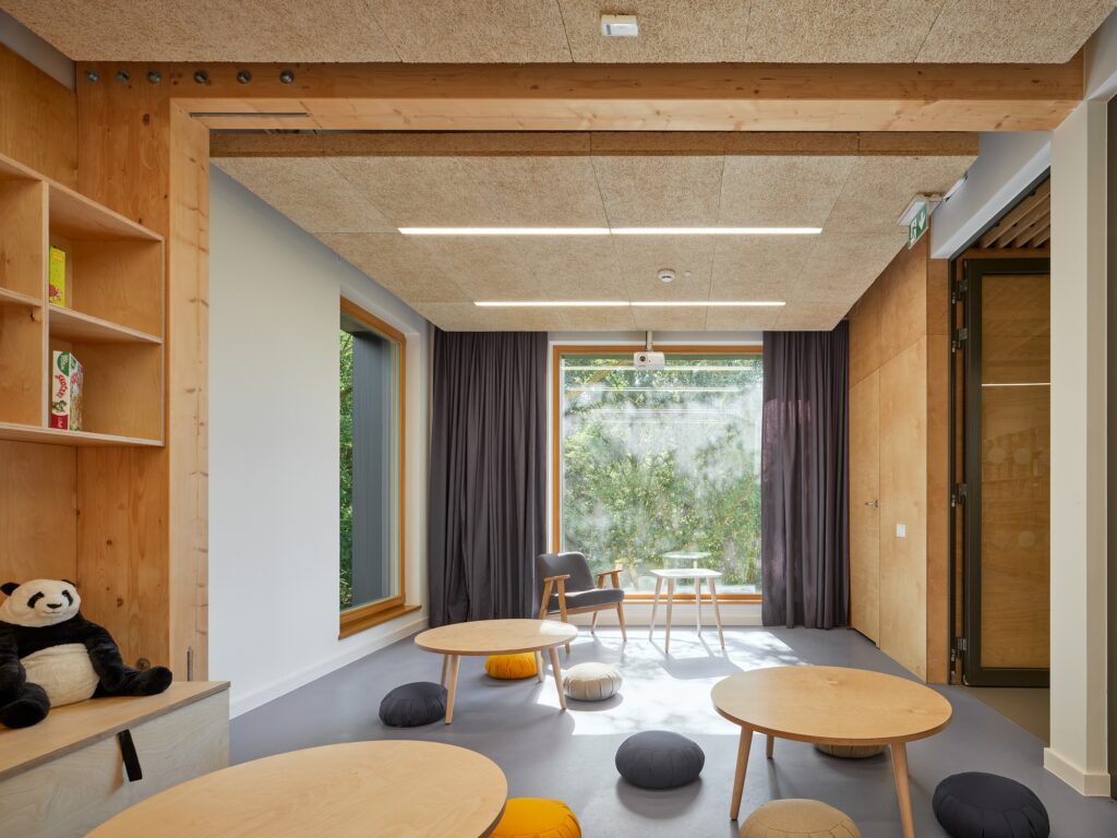 Children's reading area with low round wooden tables, floor cushions, and a large window overlooking natural foliage.