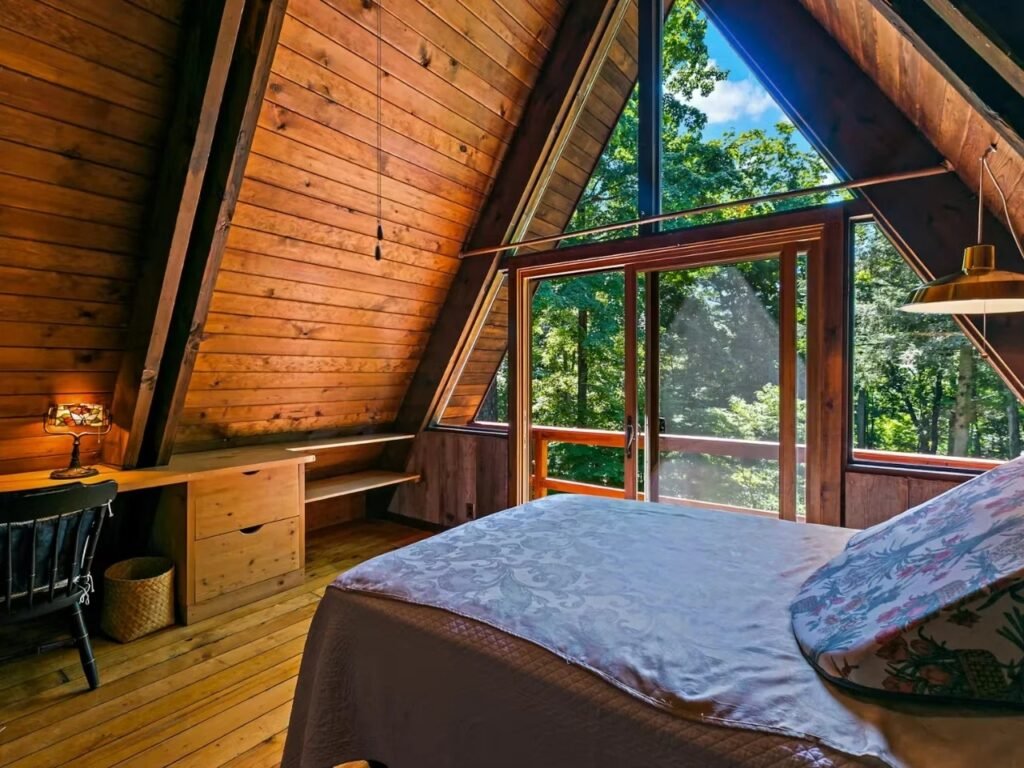 An upper-level A-frame bedroom with wood-paneled walls and ceiling beams. A bed with a patterned cover is centered. Floor-to-ceiling glass sliding doors open to a wooden balcony overlooking the forest. A built-in wooden desk with a vintage black chair and a lamp is on the left.