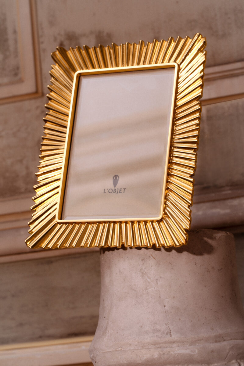 A gold ridged picture frame from the L’OBJET x Jessica McCormack collection displayed on a neutral pedestal.