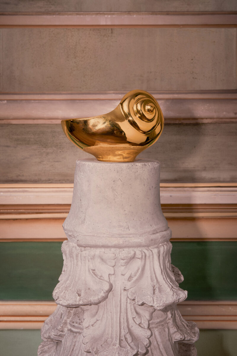A gold seashell object from the L’OBJET x Jessica McCormack collection displayed on a carved pedestal.