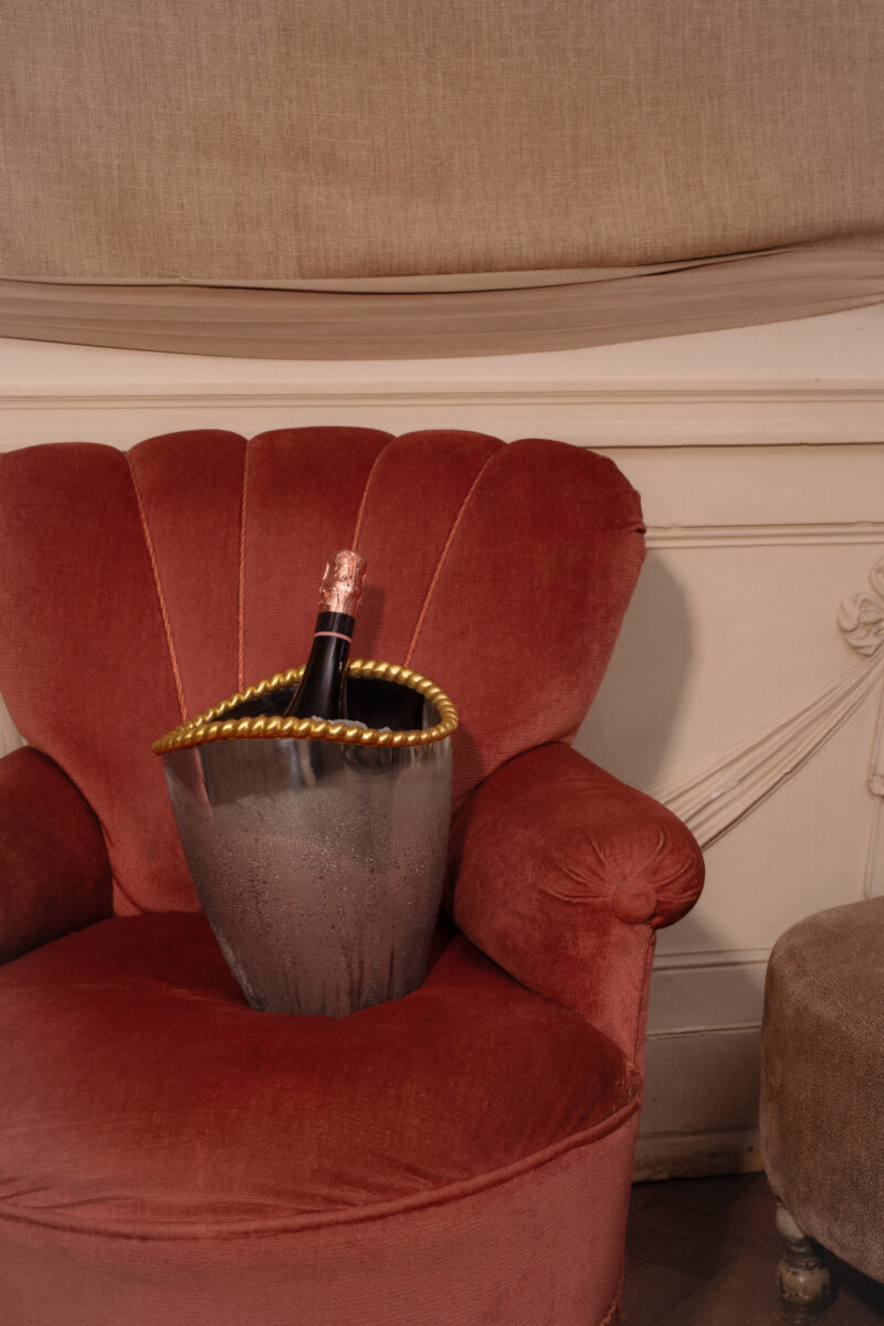 A champagne bottle in an ice bucket with a gold rope detail beside upholstered furniture.