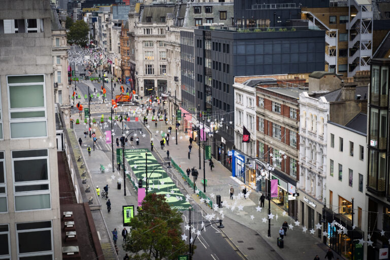 Elevated view along Oxford Street showing pedestrians, green road markings, temporary barriers and overhead star decorations.