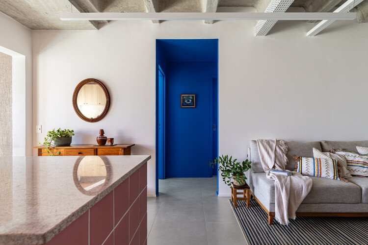 Blue entrance recess beside the pink kitchen counter and living area under an exposed ribbed ceiling