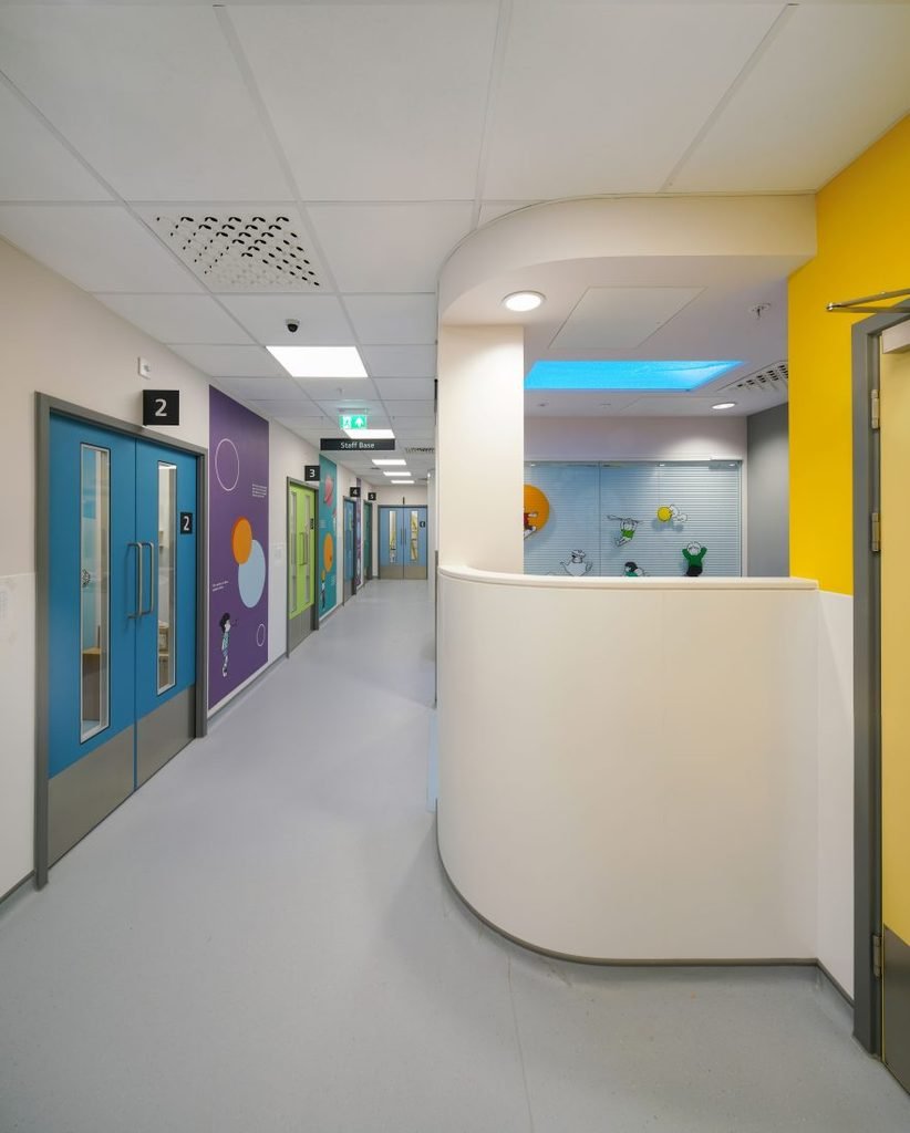 Colourful interior corridor in the Evelina Children's Day Surgery Unit, showing numbered blue doors, walls in distinct purple, green, and yellow tones, a curved white reception desk, and cartoon-style murals near the ceiling.
