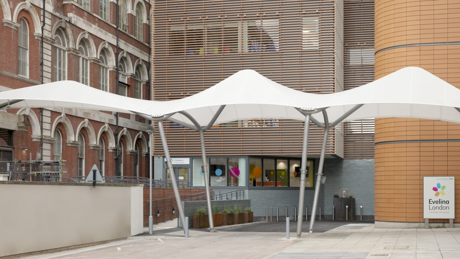 Main entrance of the Children's Day Surgery Unit at Evelina London, showing a white fabric canopy, a slatted timber-toned rainscreen fa&ccedil;ade, and the red-brick Victorian South Wing to the left.