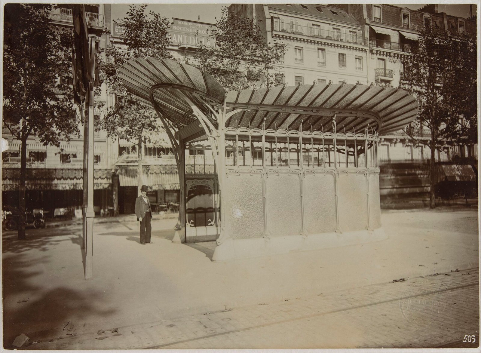 July 1900 photograph of Hector Guimard's covered Paris M&eacute;tro entrance, with a wing-shaped glazed canopy, metal enclosure and a man standing beside it.