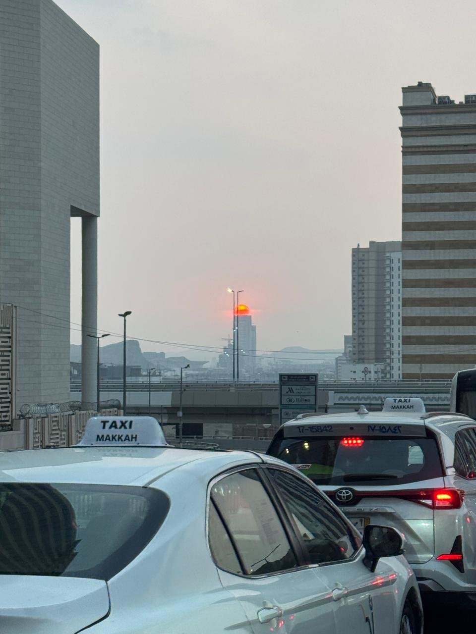 White taxis in Makkah traffic with a red sun setting behind distant skyscrapers through a hazy sky