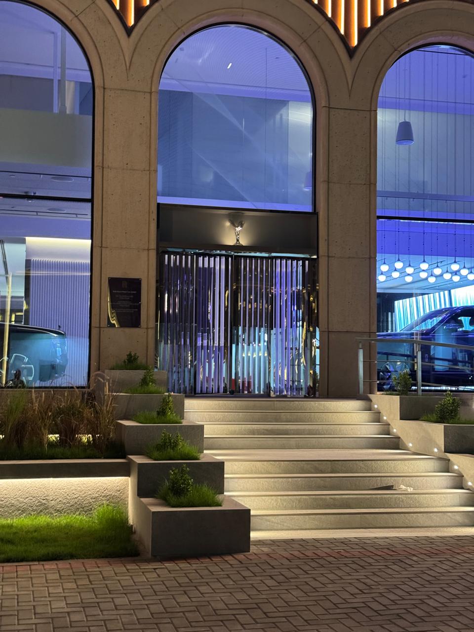 Illuminated entrance to a Rolls-Royce showroom with stone steps, terraced planters, and large arched glass doors