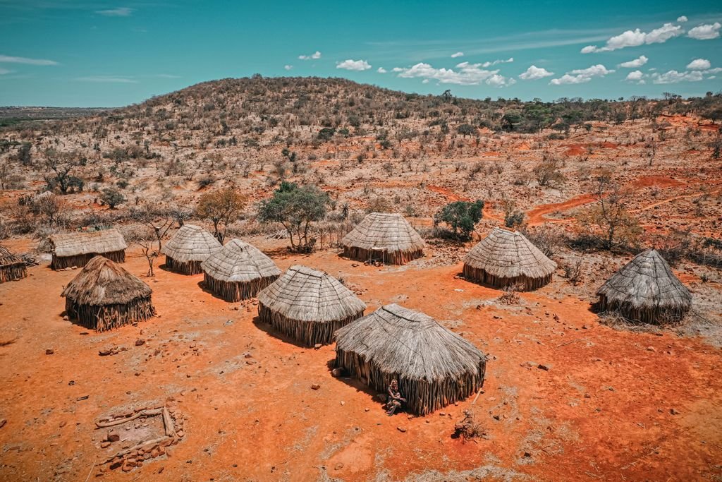Elevated view of round thatched huts arranged across red earth in a dry, sparsely wooded landscape.