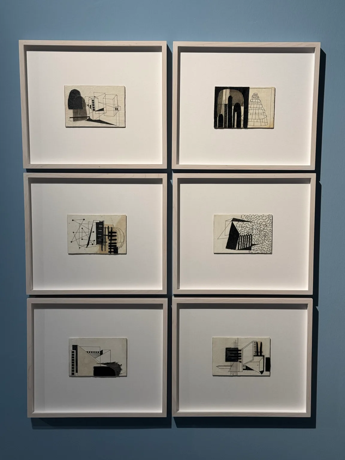 Six small framed works with architectural forms arranged in two columns on a blue gallery wall at Hayy Jameel.