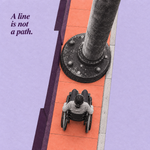 A wheelchair user faces a large pole blocking a narrow sidewalk, with the words &ldquo;A line is not a path.&rdquo; | مستخدم كرسي متحرك أمام عمود يسد رصيفًا ضيقًا، مع عبارة &laquo;الخط ليس مسارًا&raquo;.