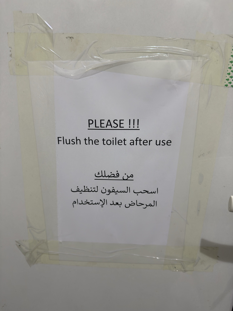A printed Arabic and English notice asking users to flush the toilet is attached to a white surface with strips of tape.