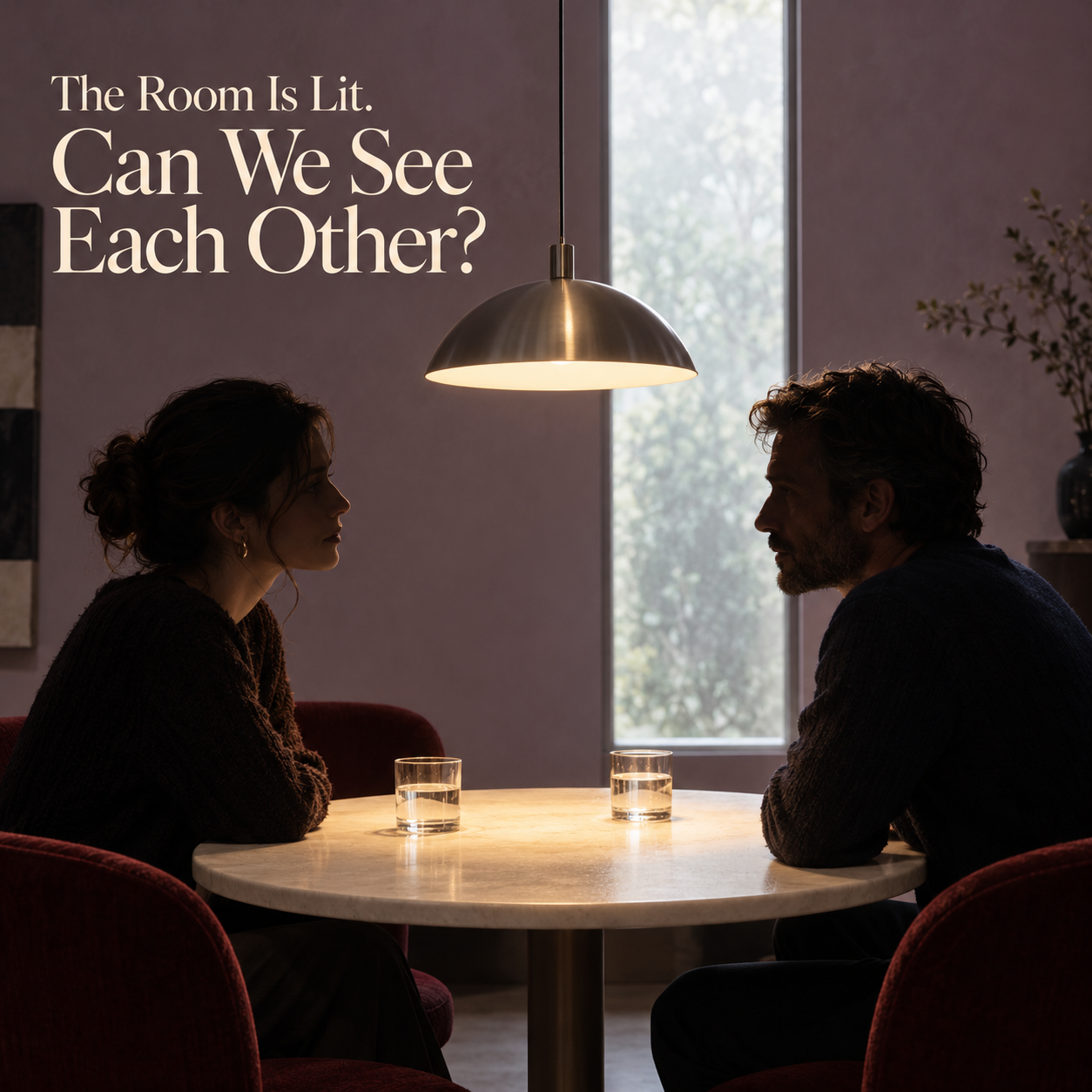 Two people face each other across a lit round table while their faces remain in shadow beside a bright window.