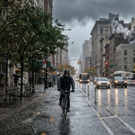 A cyclist rides along a designated cycle lane on a wet city street beneath dark, gloomy skies