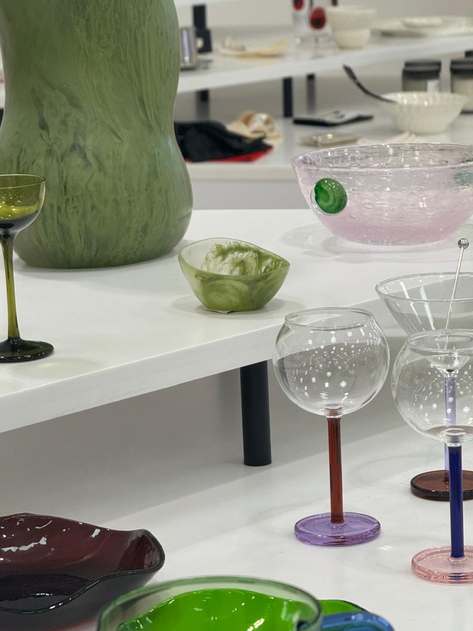 Colourful glassware on display, including a large green vase, pink bubbly bowl, and clear glasses with coloured stems