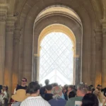 Visitors in a vaulted stone passage at the Louvre Museum in Paris, with the glass pyramid visible ahead.