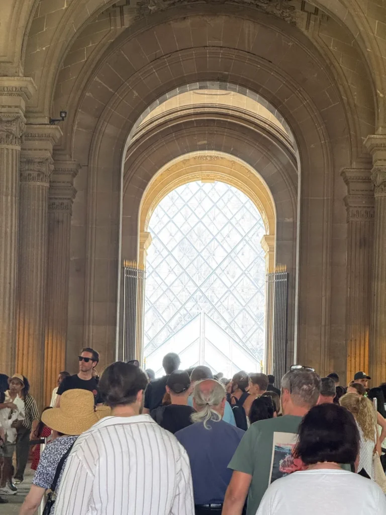 Visitors in a vaulted stone passage at the Louvre Museum in Paris, with the glass pyramid visible ahead.