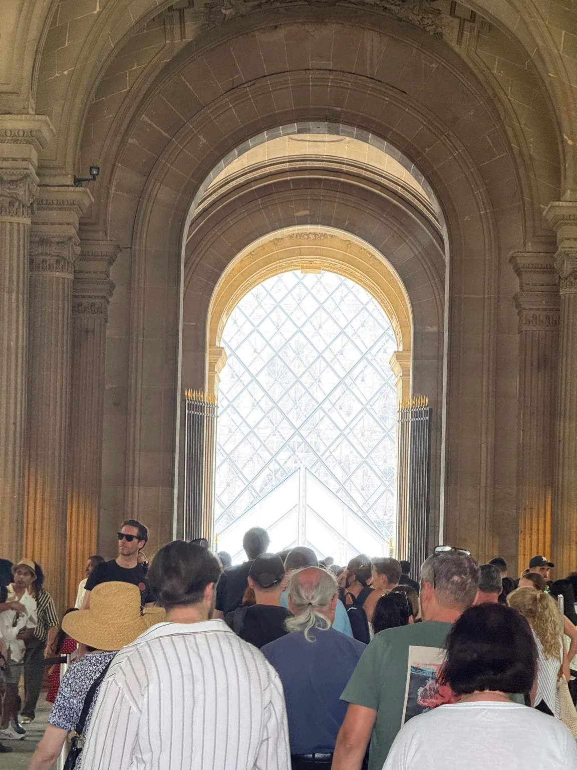 Visitors in a vaulted stone passage at the Louvre Museum in Paris, with the glass pyramid visible ahead.