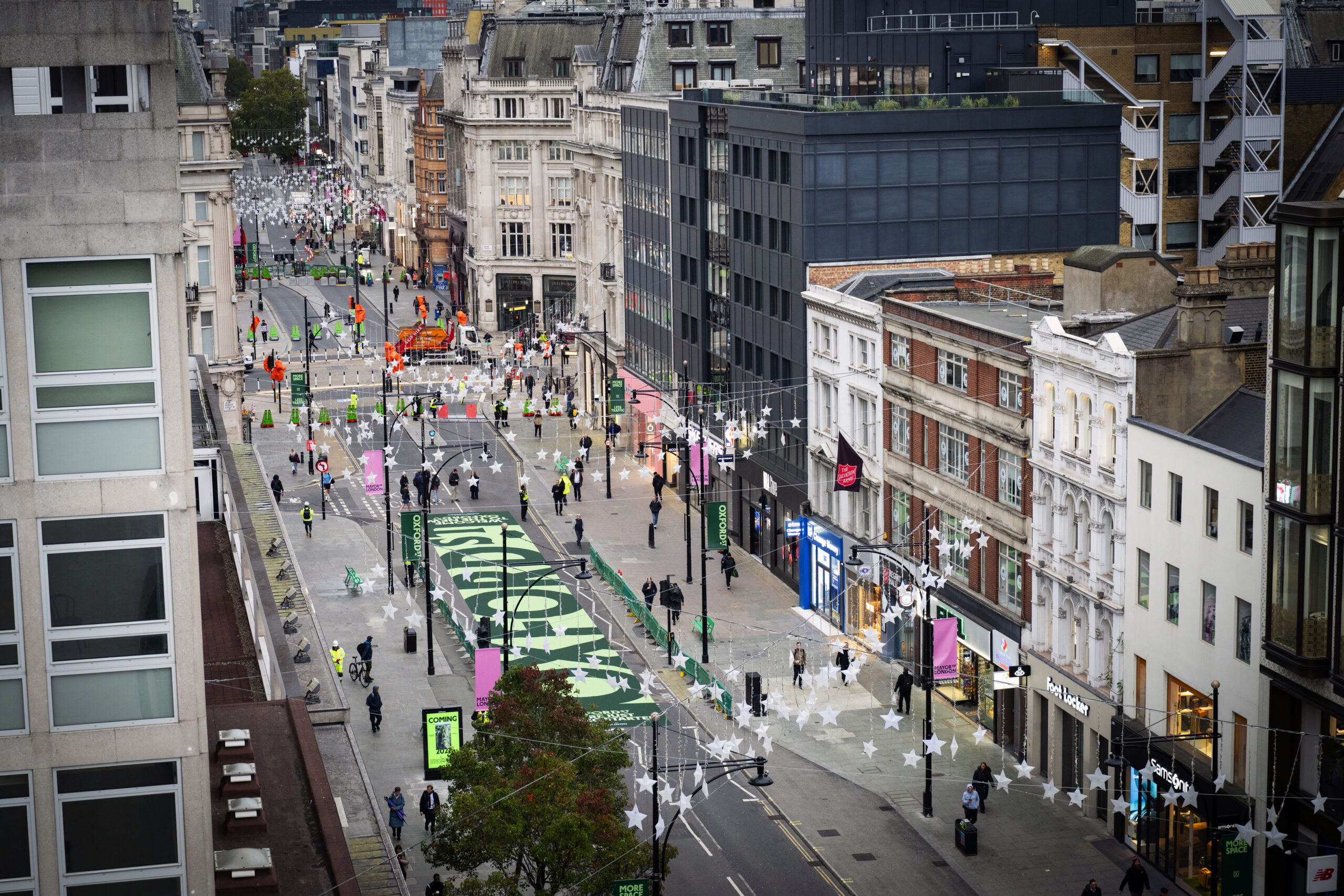 Elevated view along Oxford Street showing pedestrians, green road markings, temporary barriers and overhead star decorations.